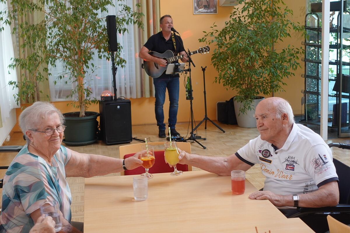 Die beiden Bewohner:innen Ilse Kratzer und Johann Grandits stoßen mit einem bunten Cocktail in der Hand an und genießen die wundervolle Musik von Mitarbeiter Christian Loier.
